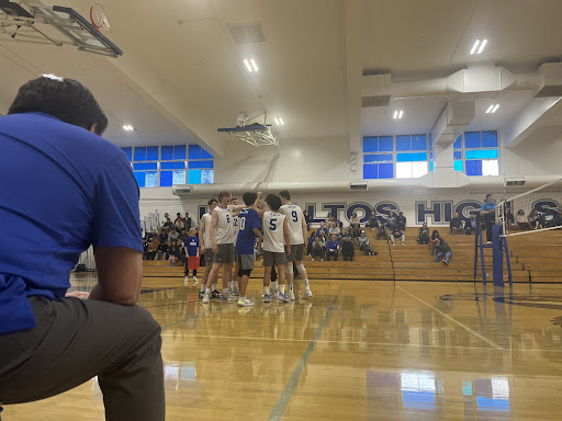 The varsity boys volleyball team huddles for a cheer before their last game, as their head coach looks on.