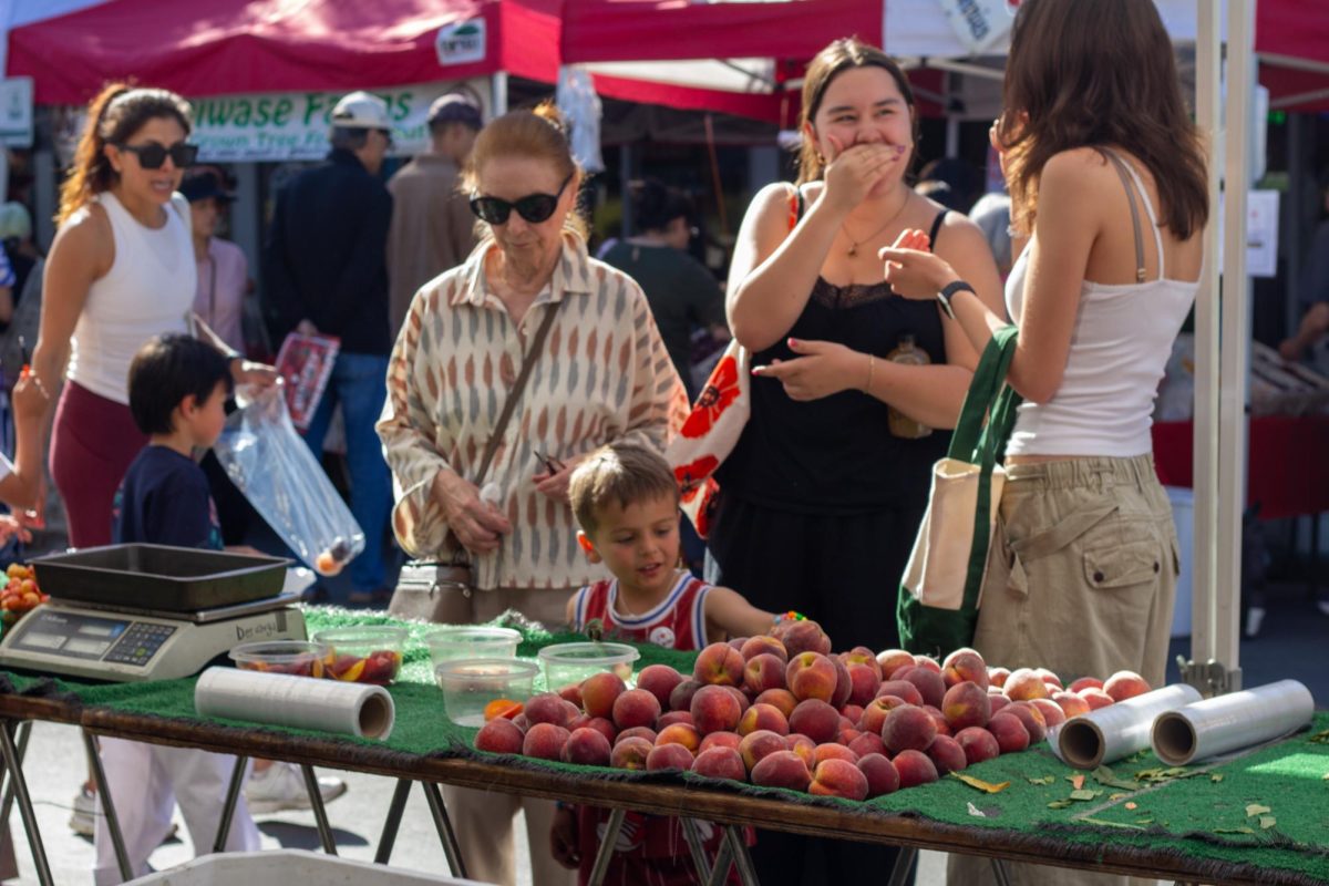 A child reaches toward a display of fresh peaches as shoppers stroll through the farmers’ market. The market’s lively atmosphere invites families, friends, and local vendors each Thursday. 